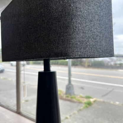 Black lamp with a textured shade in front of a window with a blurred street view.