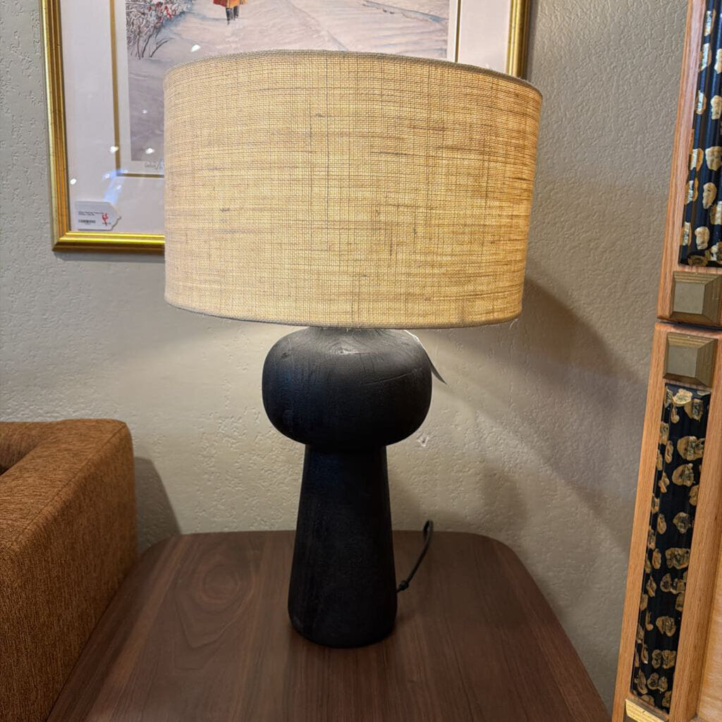 Table lamp with a woven shade on a wooden surface, with a textured wall and framed picture in the background.