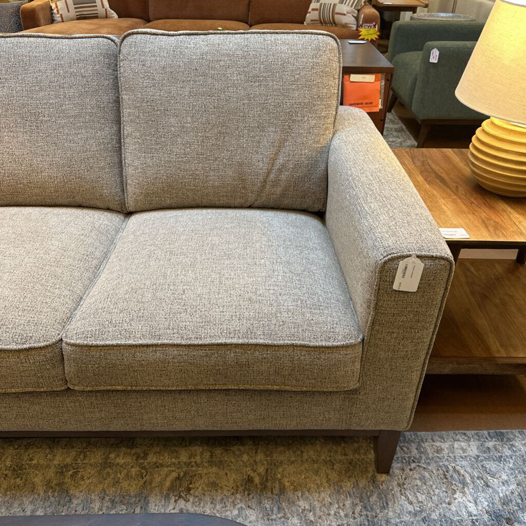 Beige sofa in a furniture store setting with a lamp and other furniture items in the background.
