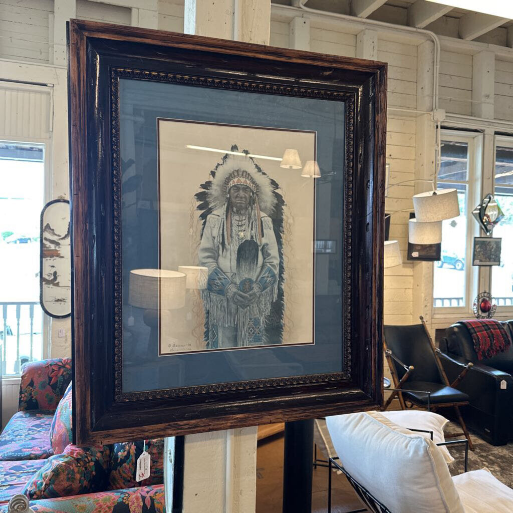 Framed artwork of a Native American figure in a headdress on a stand in a room with furniture.