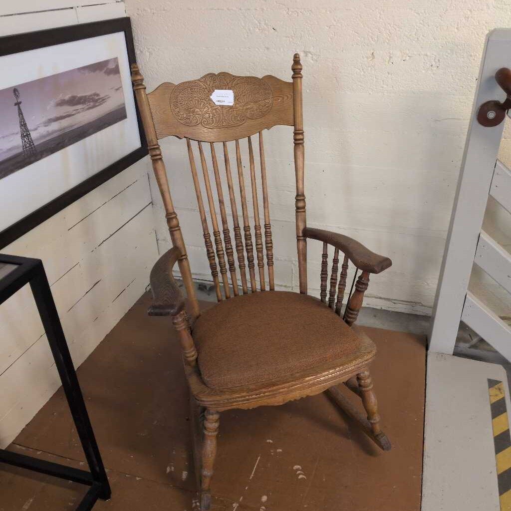 Wooden rocking chair with a brown cushion on a wooden floor, with a framed picture and ladder in the background.