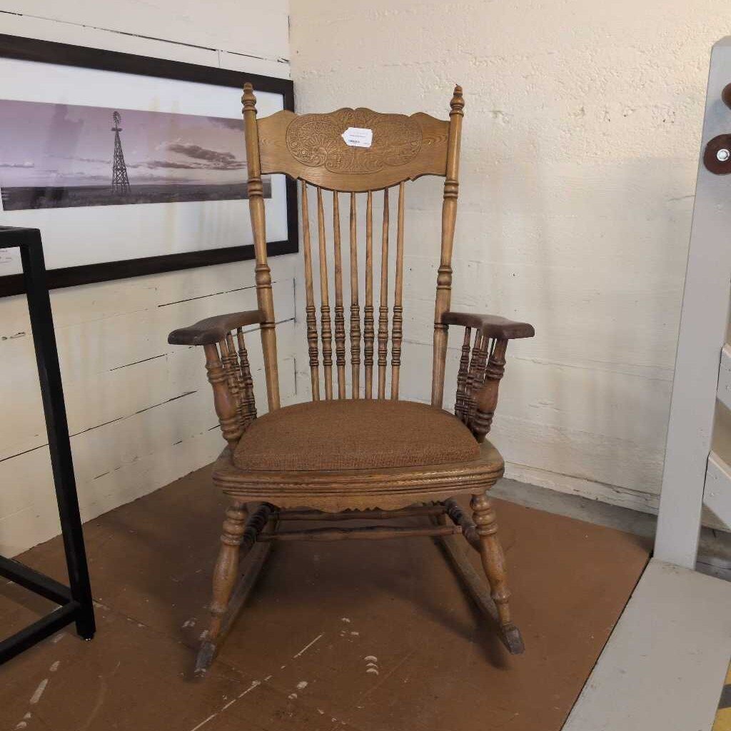 Wooden rocking chair with a brown cushion on a floor with a white wall and framed picture in the background.