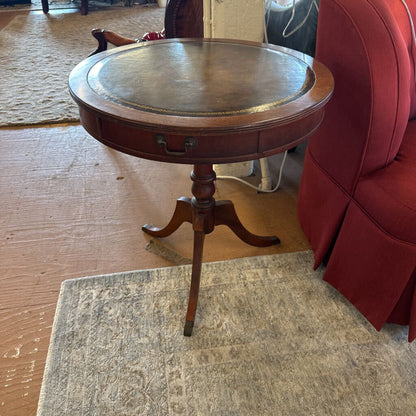 Round wooden side table with a drawer in a room with a red chair and wooden floor.