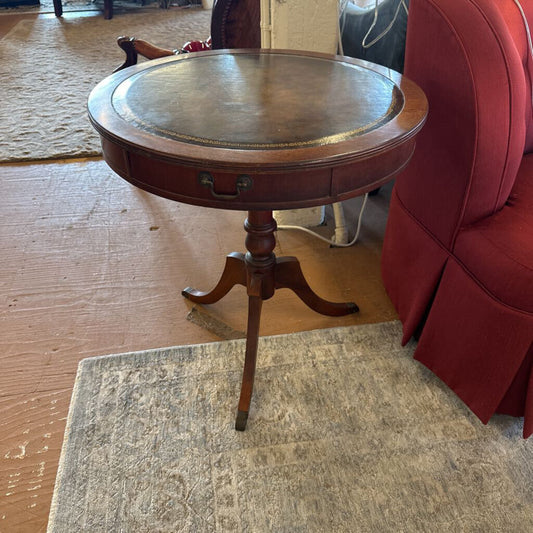 Round wooden side table with a drawer in a room with a red chair and wooden floor.