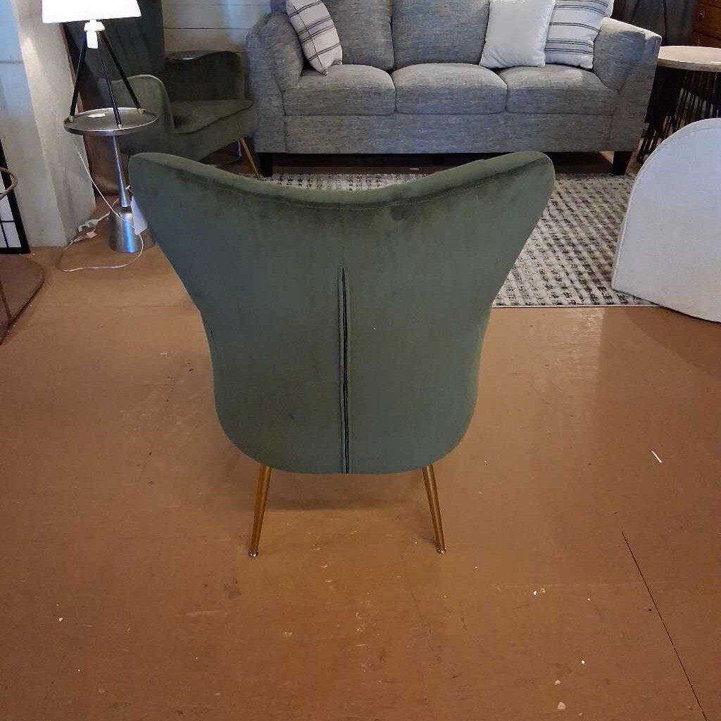 Green chair with gold legs on a brown floor, with a gray sofa and lamp in the background.