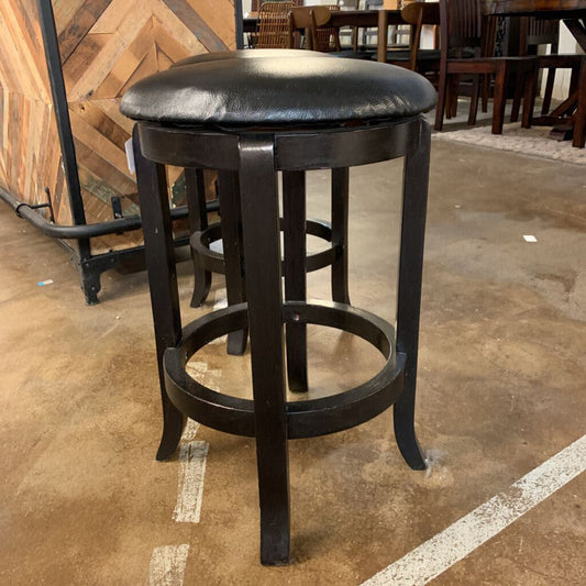 Black leather bar stool on a concrete floor with wooden walls and furniture in the background.