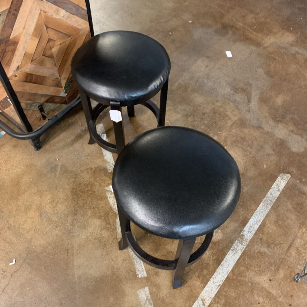 Two black bar stools on a concrete floor with a wooden shelf in the background.