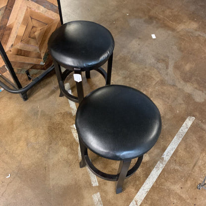 Two black bar stools on a concrete floor with a wooden shelf in the background.