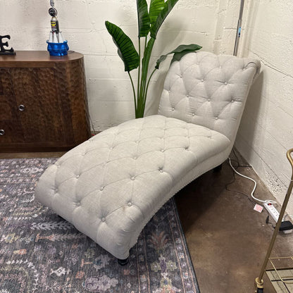 White tufted chaise lounge chair in a room with a plant and wooden cabinet.