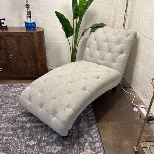 White tufted chaise lounge chair in a room with a plant and wooden cabinet.