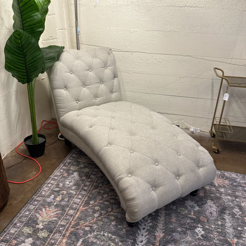 Light gray tufted chaise lounge on a patterned rug with a plant and cart in the background.