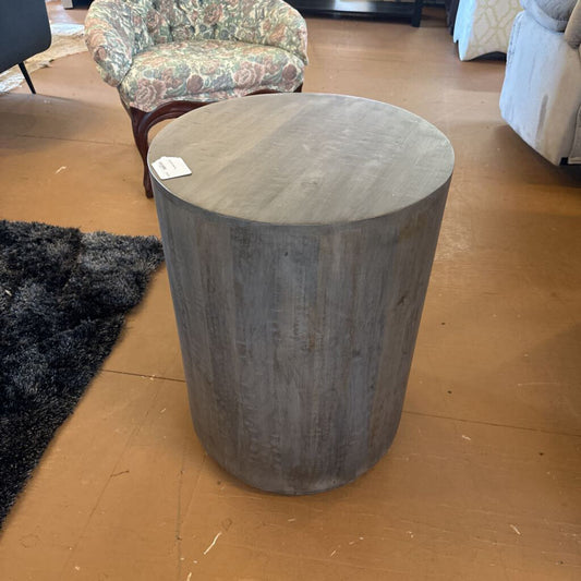 Round wooden stool on a wooden floor with a chair and rug in the background