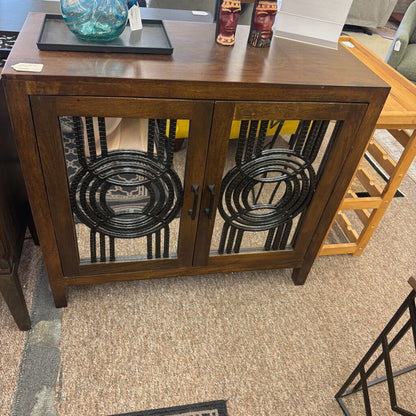 Wooden cabinet with glass doors featuring decorative metalwork on a carpeted floor.