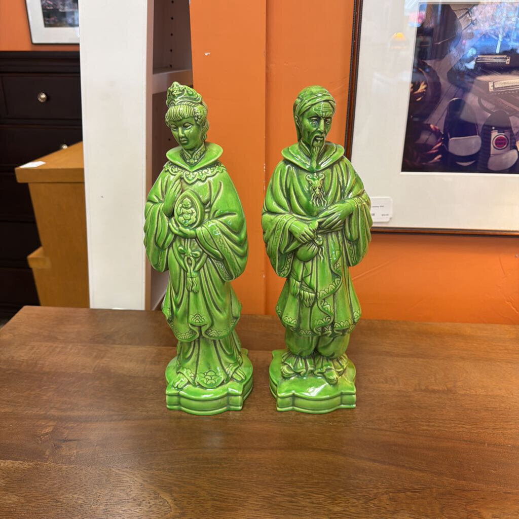 Two green ceramic statues on a wooden surface with an orange wall in the background.