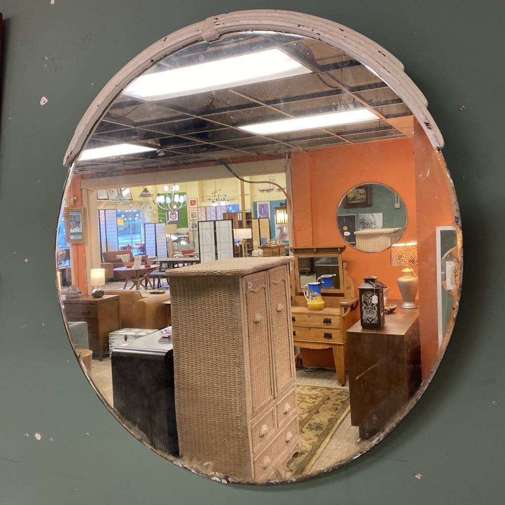 A large, round vintage wall mirror with a white distressed frame, reflecting a furniture showroom filled with wicker cabinets and wooden dressers