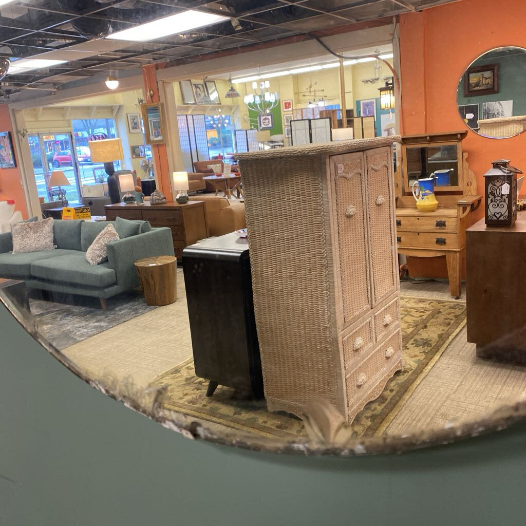 A large, round vintage wall mirror with a white distressed frame, reflecting a furniture showroom filled with wicker cabinets and wooden dressers