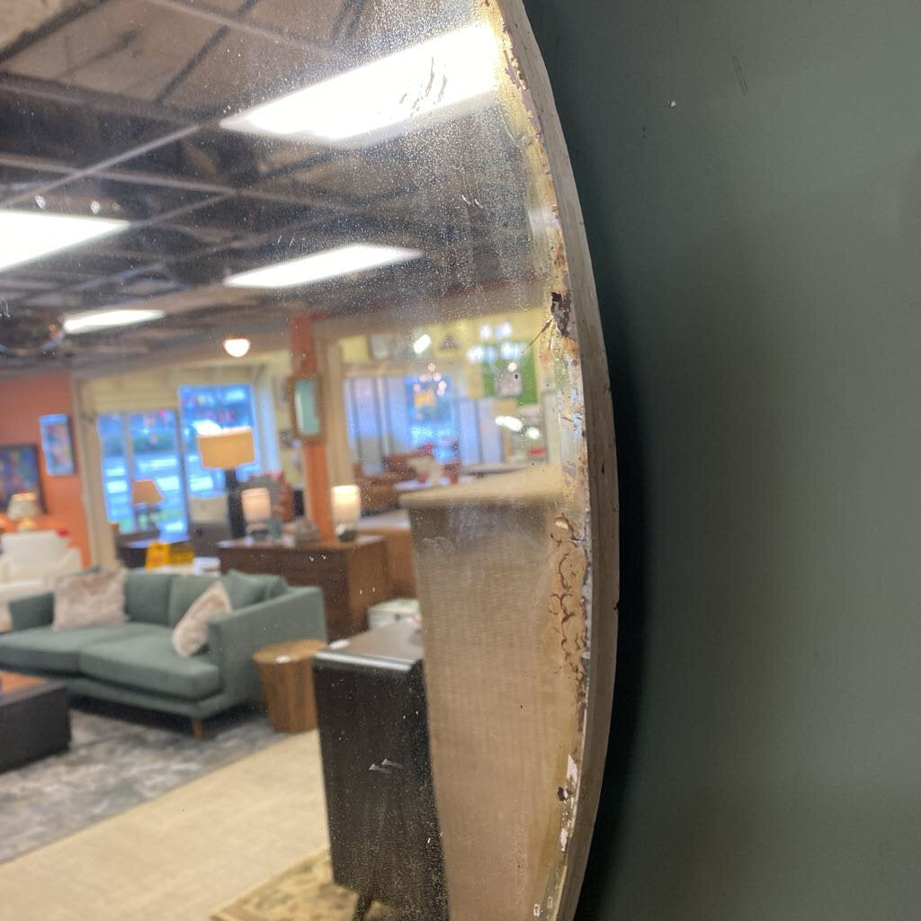 A large, round vintage wall mirror with a white distressed frame, reflecting a furniture showroom filled with wicker cabinets and wooden dressers