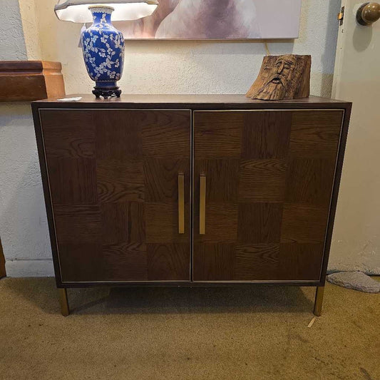 Wooden cabinet with two doors on a carpeted floor, with a lamp and decorative piece in the background.