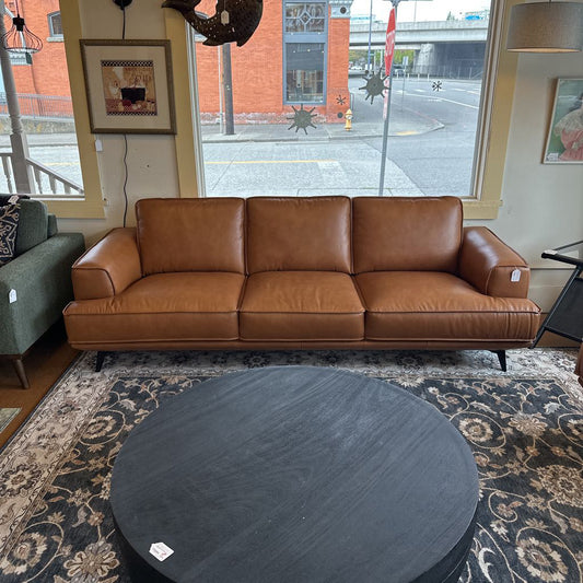 Brown leather sofa in a room with a round dark coffee table and a window view.