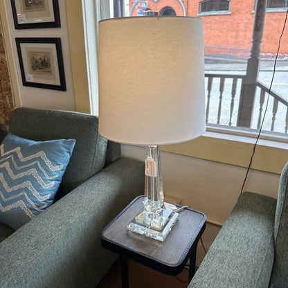 Table lamp with beige shade on a side table next to a gray sofa with a blue pillow.