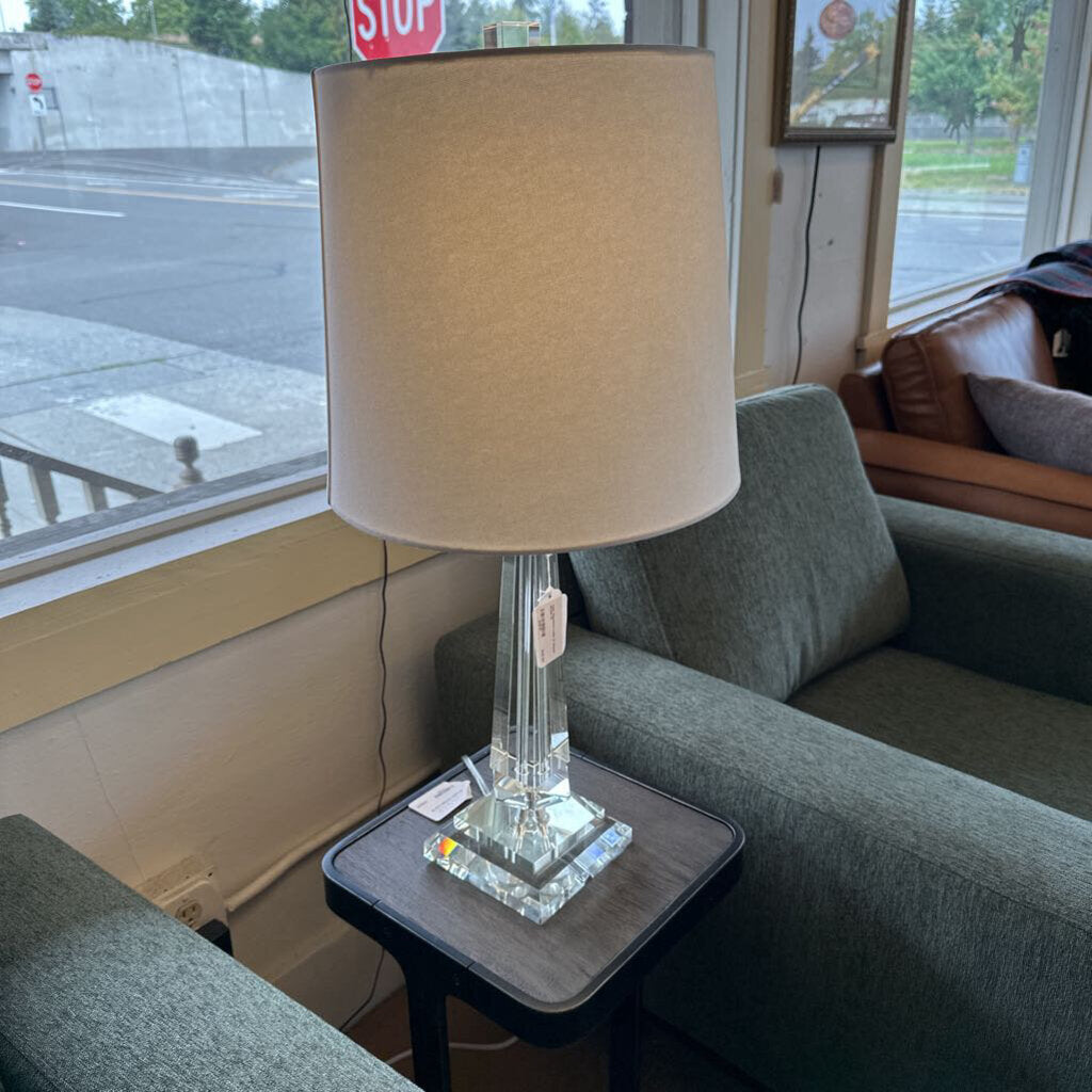 Lamp with beige shade on a side table in a room with a couch and window view.