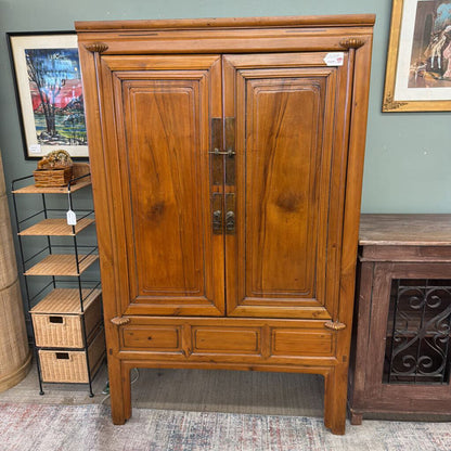 Wooden armoire with two doors in a room setting with shelves and artwork.