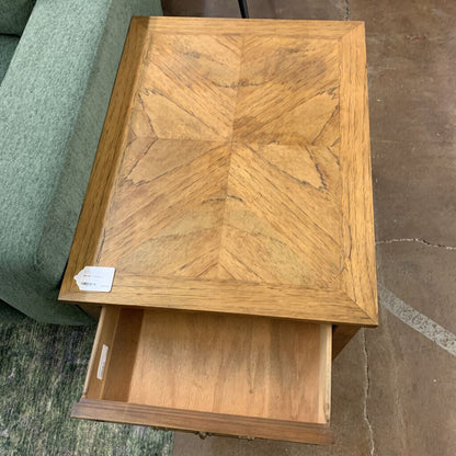 Wooden side table with chevron pattern and open drawer on a carpeted floor.
