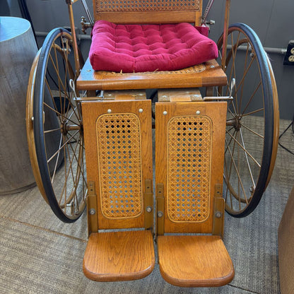 Vintage wooden wheelchair with wicker backrest and pink cushion on a carpeted floor.