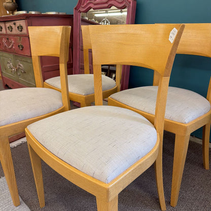 Four wooden chairs with beige cushioned seats in a room with a mirror and furniture.