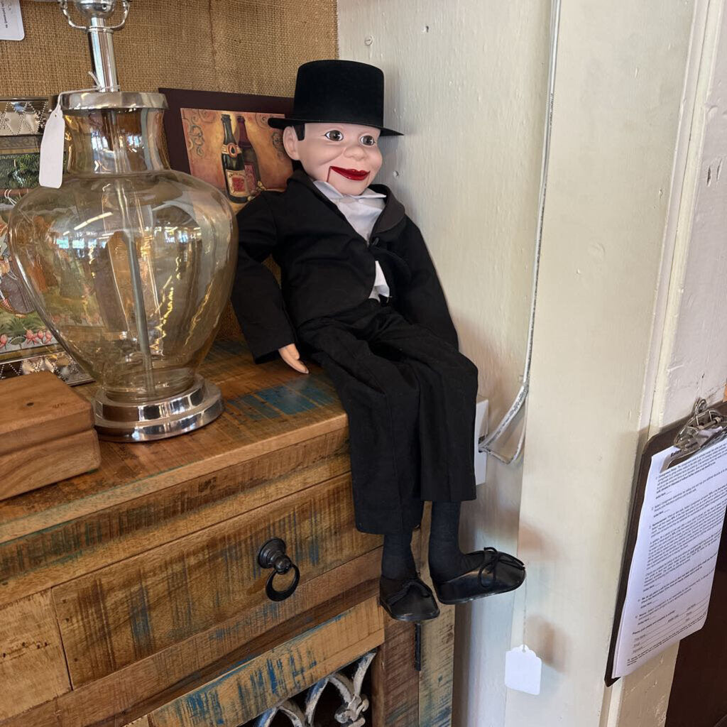 Vintage-style doll in a black outfit and hat on a wooden dresser with a glass lamp.