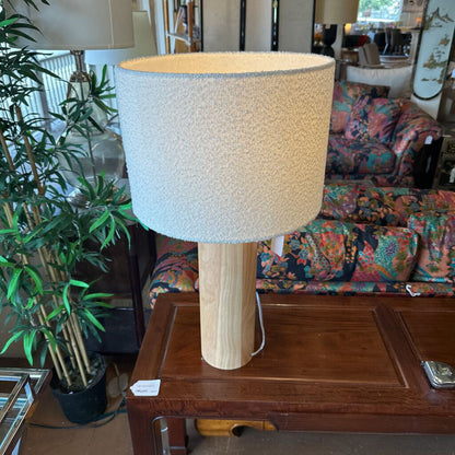 Table lamp with a textured beige shade on a wooden table in a room with furniture and plants.