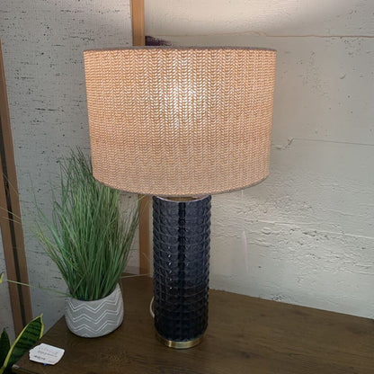 Table lamp with textured beige shade and dark base on a wooden surface, with a plant in the background.