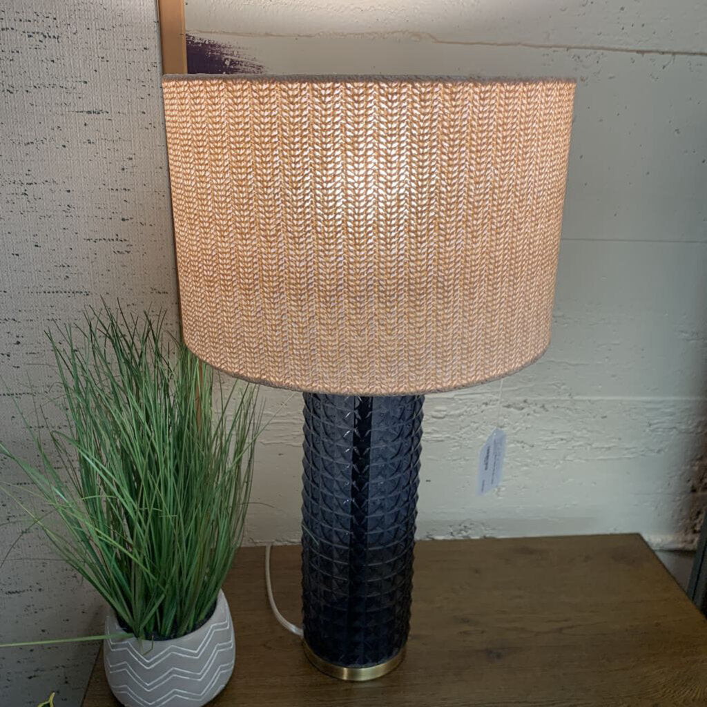 Table lamp with woven shade and textured black base on a wooden surface, next to a plant.