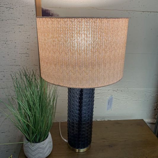 Table lamp with woven shade and textured black base on a wooden surface, next to a plant.