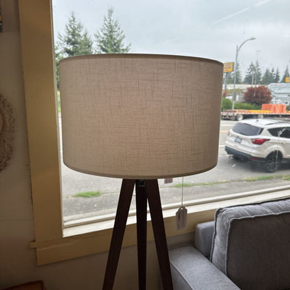 Floor lamp with a beige shade and wooden base in a room with a window showing a street view.