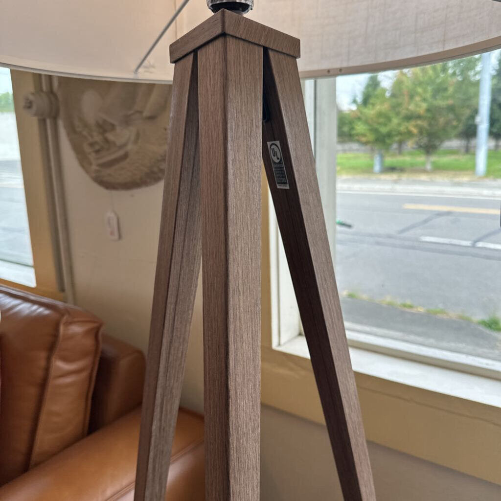 Wooden lamp stand with a beige lampshade in a room with a window and brown sofa.