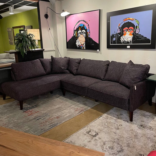 A dark grey, L-shaped fabric sectional sofa with tapered wooden legs, styled on a patterned rug beneath two pop-art paintings of chimpanzees wearing headphones.