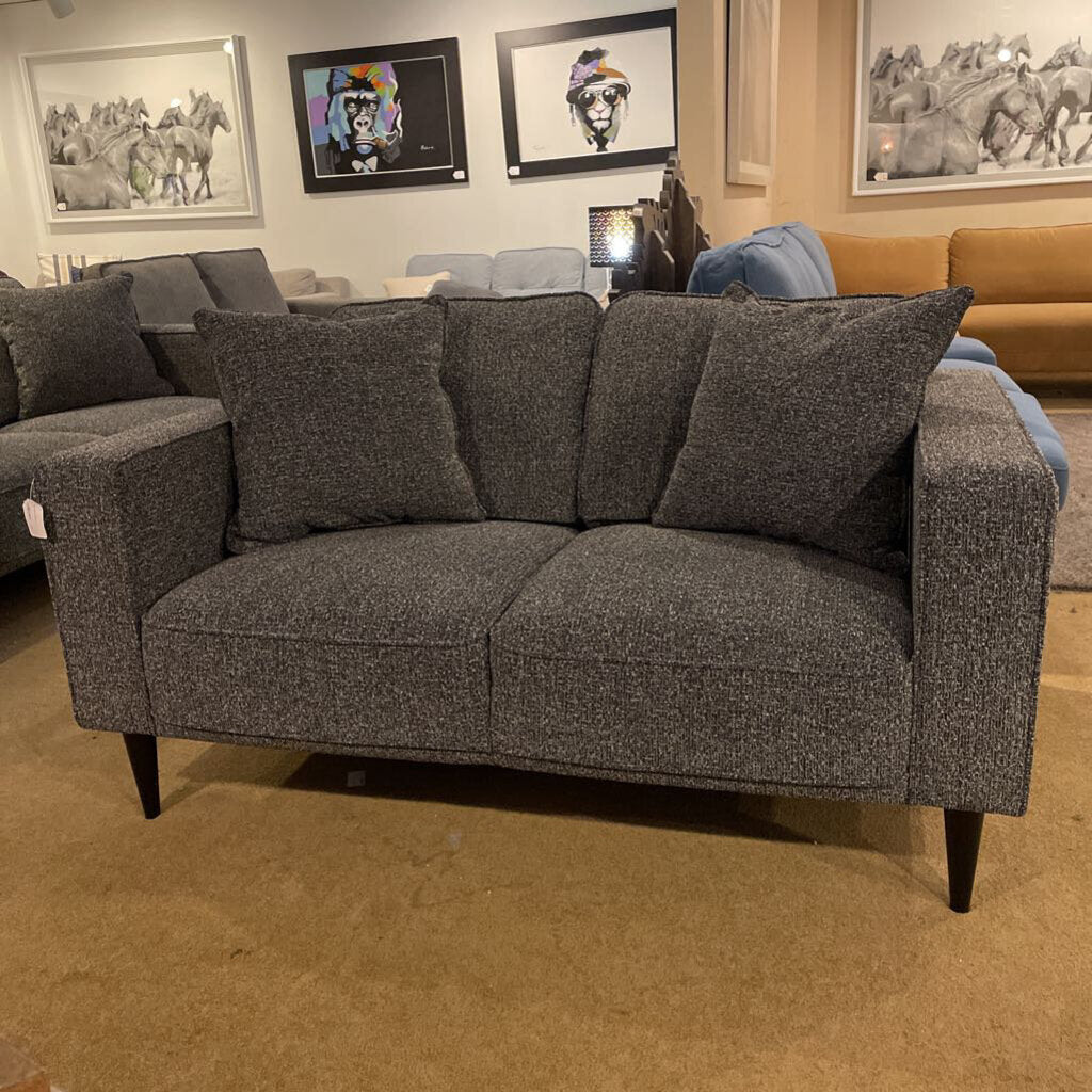 A contemporary charcoal grey two-seater loveseat with square arms and tapered black legs, shown in a furniture showroom.