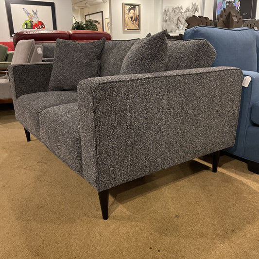 A contemporary charcoal grey two-seater loveseat with square arms and tapered black legs, shown in a furniture showroom.