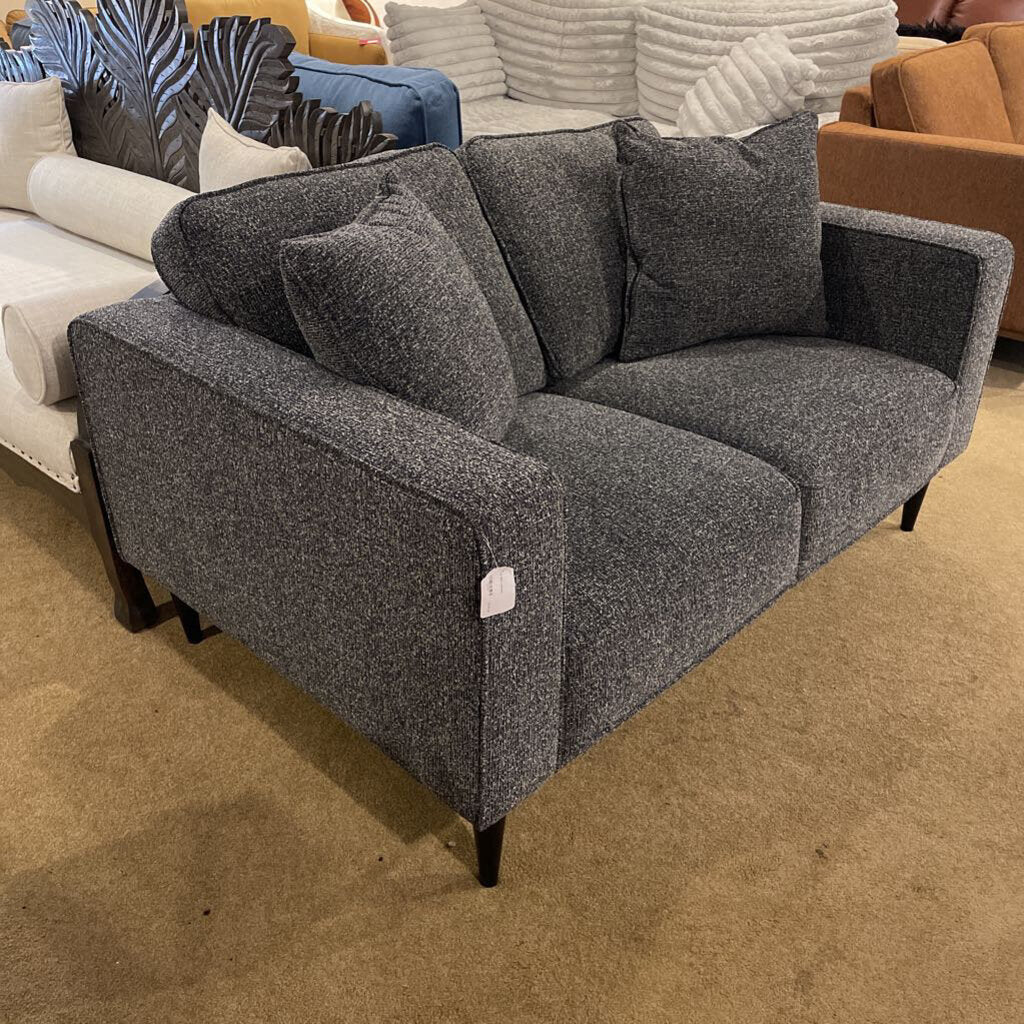 A contemporary charcoal grey two-seater loveseat with square arms and tapered black legs, shown in a furniture showroom.