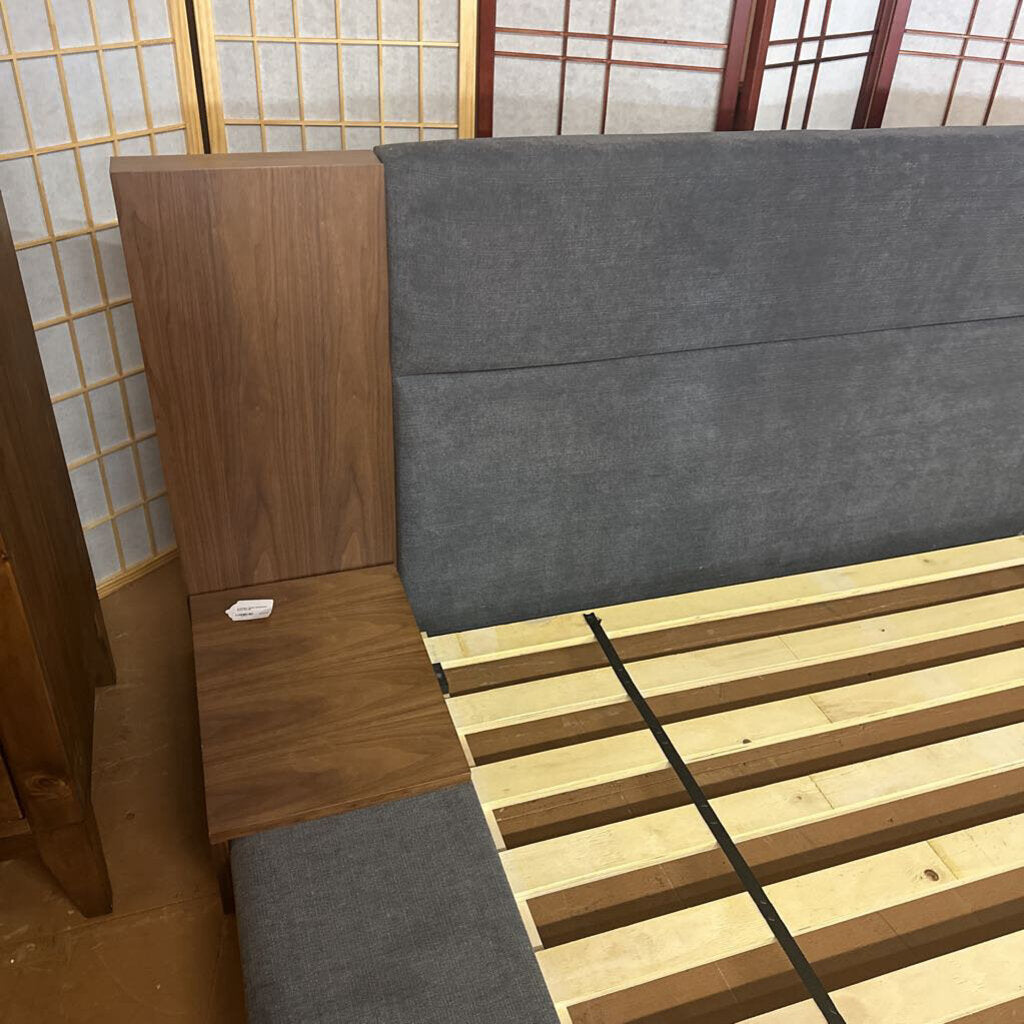 Gray upholstered bed with wooden slats and a wooden headboard against a wall with a grid pattern.