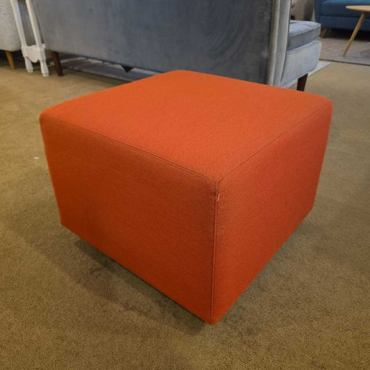 Orange cube-shaped ottoman on a carpeted floor with a blurred background