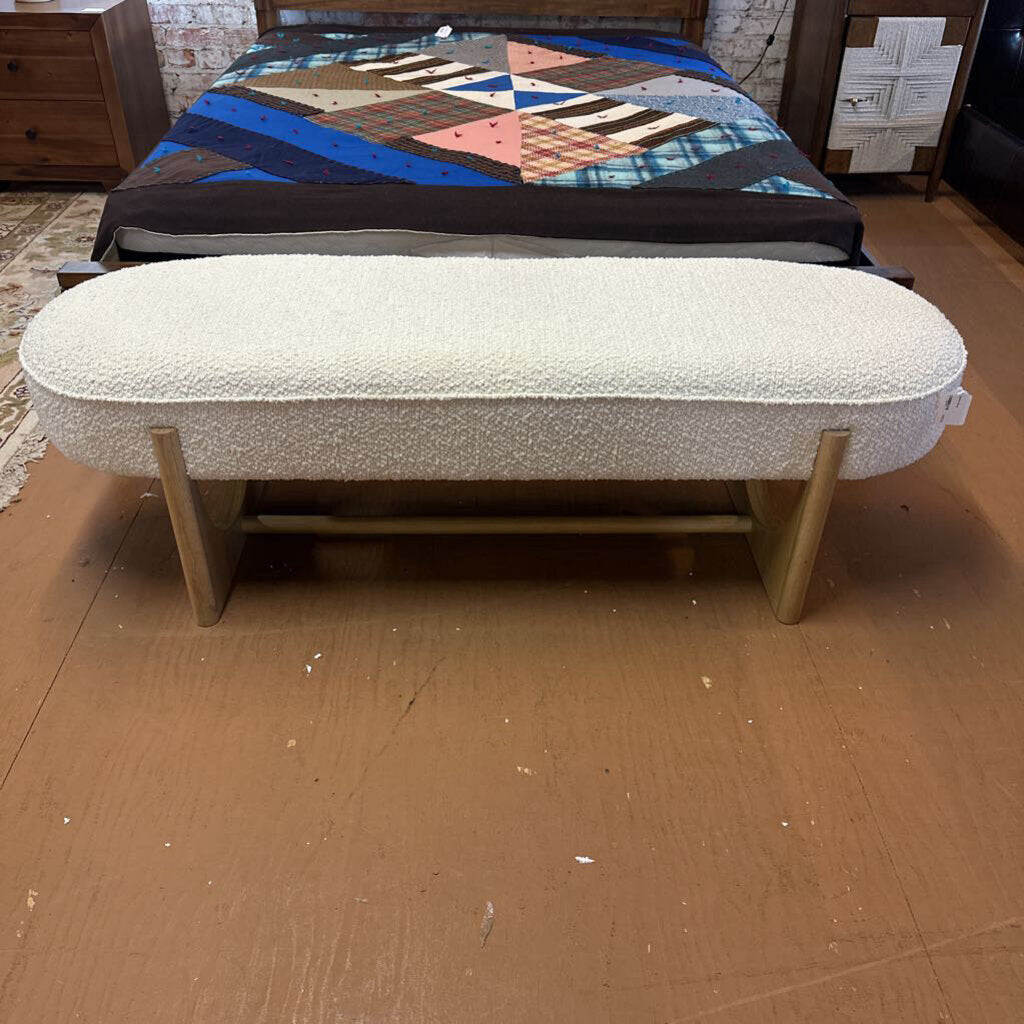 White cushioned bench with wooden legs in front of a bed with a quilt.