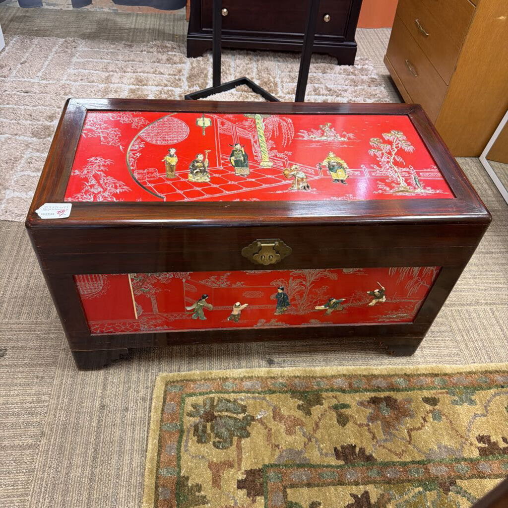 Decorative wooden chest with red lid featuring gold and green figures on a carpeted floor.