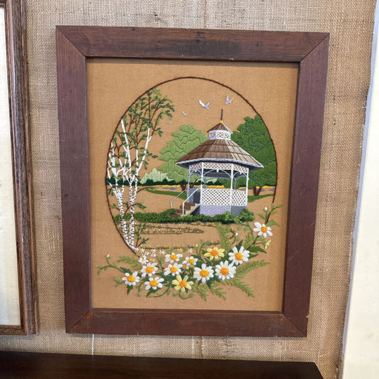 Framed needlepoint of a garden scene with a gazebo and flowers on a textured fabric background.