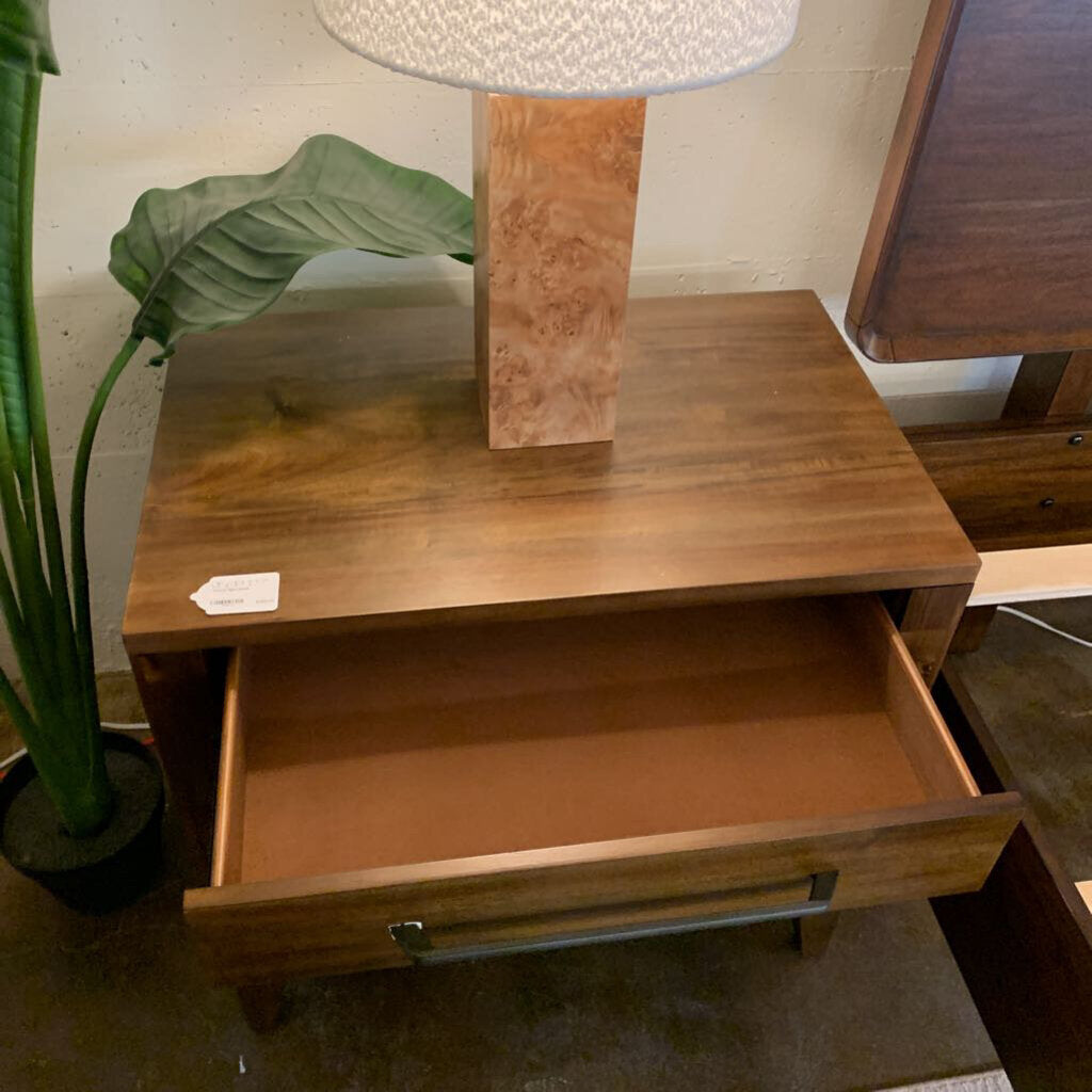 Wooden nightstand with open drawer, lamp, and plant in the background