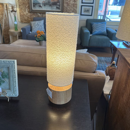 Tall cylindrical lamp with woven design on a dark surface in a living room setting.