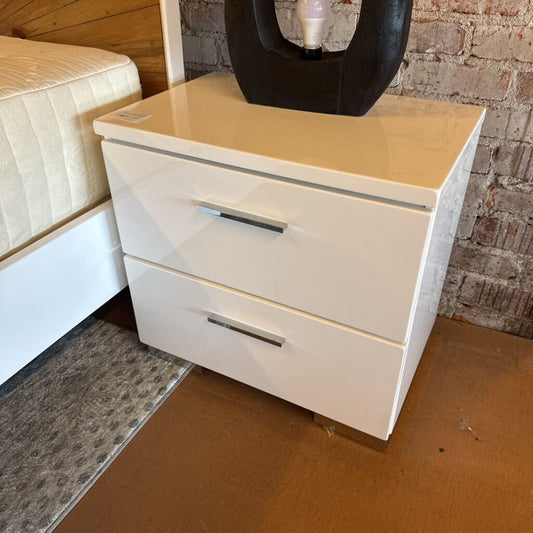 White nightstand with two drawers against a brick wall.