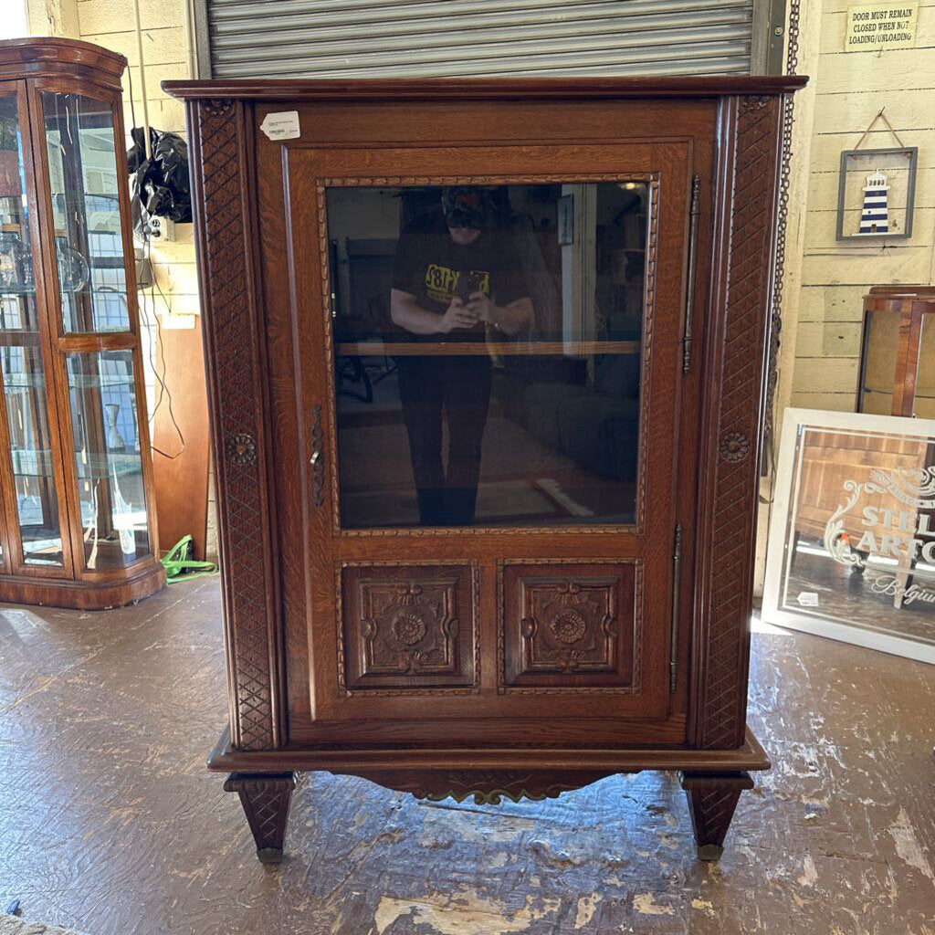 Wooden cabinet with glass door in a room with tiled walls and another cabinet in the background.