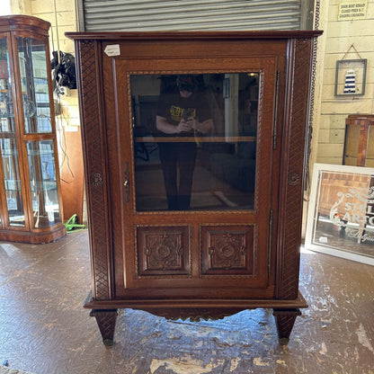Wooden cabinet with glass door in a room with tiled walls and another cabinet in the background.